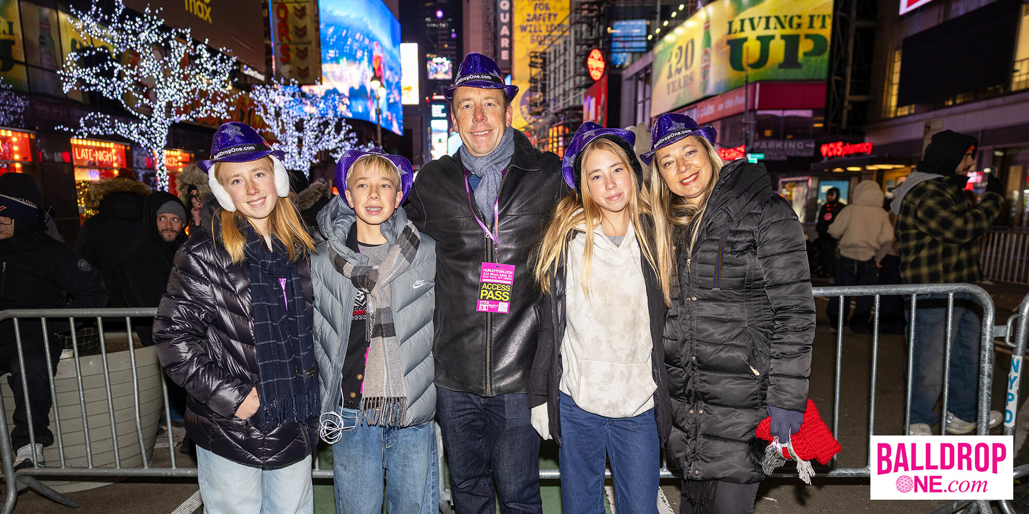 Group of five people—two adult women, one adult man, and two children—posing in winter jackets and purple sequin hats labeled BallDropOne.com on a city street at night with festive lights and crowd barriers.